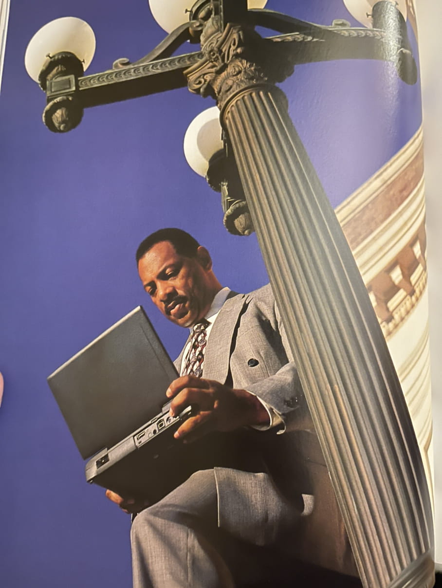 Black man in sharp suit using computer standingg against lightpole deep blue sky